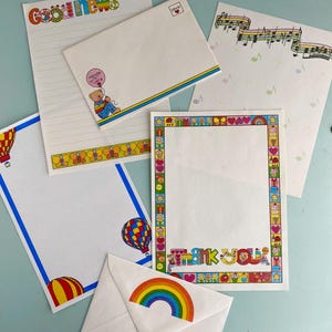 May include: A collection of stationery items, including lined paper and envelopes, with colorful illustrations. The paper features designs such as hot air balloons, musical notes, and the words "Good News" and "Thank You". The envelopes have rainbow and teddy bear graphics.