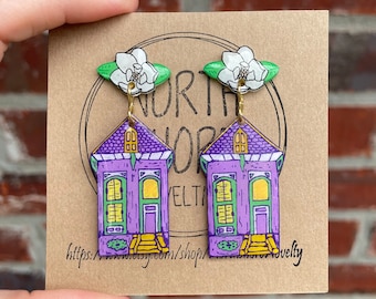 May include: A pair of purple house-shaped earrings with gold-colored hooks. Each earring features a white magnolia flower with green leaves at the top. The earrings are on a brown card with the text "NORTH SHORE NOVELTY".