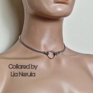 May include: A silver chain choker necklace with a large silver ring in the center. The text "Collared by Lia Nerula" is printed below the necklace.