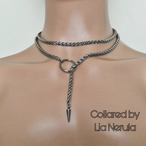 May include: A silver chain choker necklace with a ring and a pointed end. The necklace is layered and has a braided chain design. The text "Collared by Lia Nerula" is visible at the bottom of the image.