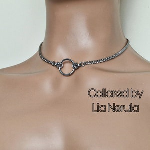 May include: A silver chain choker necklace with a large silver ring in the center. The text "Collared by Lia Nerula" is visible below the necklace.