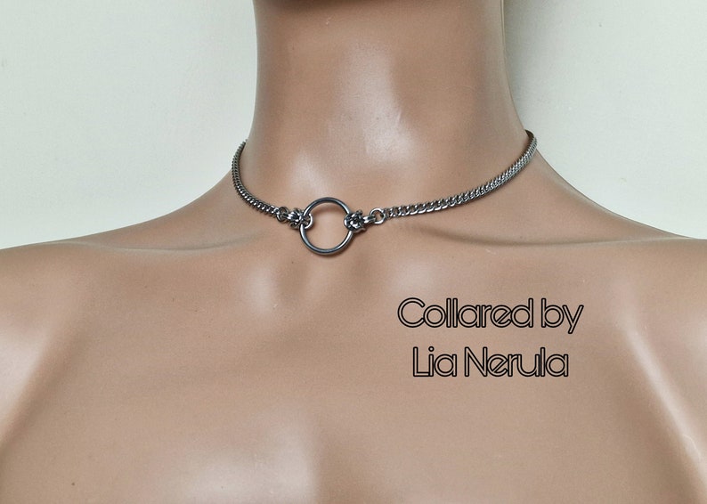 May include: A silver chain choker necklace with a large silver ring in the center. The text "Collared by Lia Nerula" is visible below the necklace.