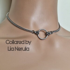 May include: A silver choker necklace with a large, round ring in the center. The necklace is made of a chain link design. The text "Collared by Lia Nerula" is printed below the necklace.