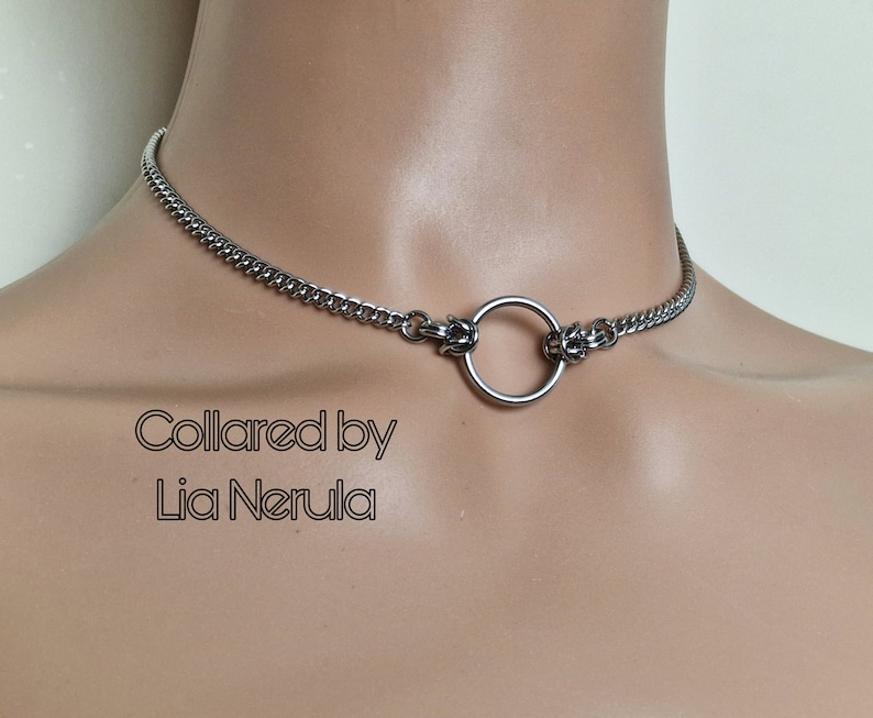 May include: A silver choker necklace with a large, round ring in the center. The necklace is made of a chain link design. The text "Collared by Lia Nerula" is printed below the necklace.