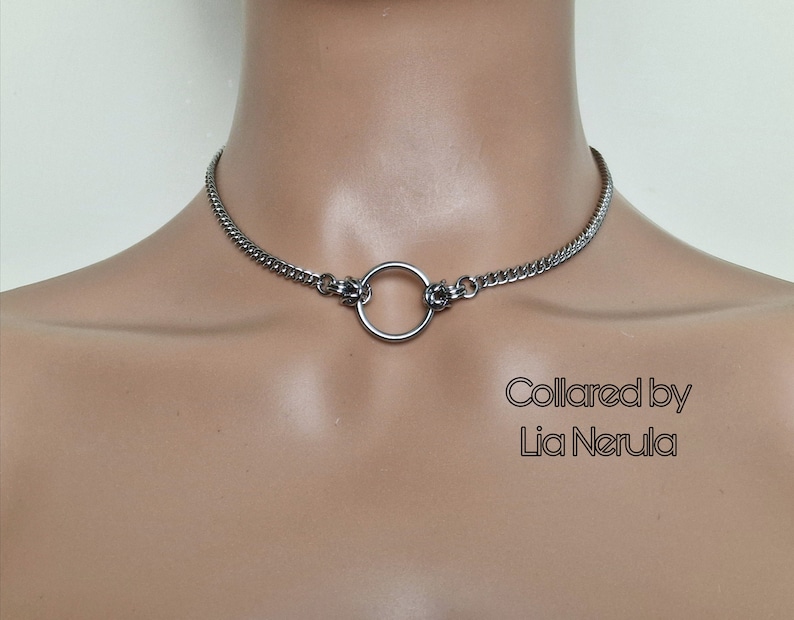 May include: A silver chain choker necklace with a large silver ring attached. The text "Collared by Lia Nerula" is printed below the necklace.