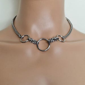 May include: A silver chain choker necklace with three silver rings. The center ring is larger than the two side rings.