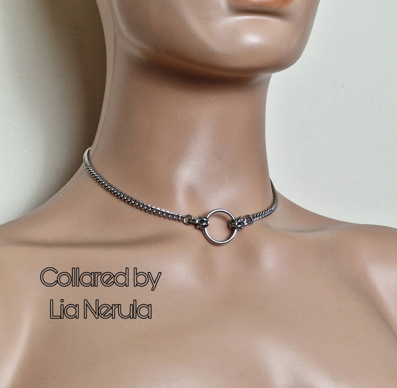 May include: A silver chain choker necklace with a large silver ring in the center. The text "Collared by Lia Nerula" is printed below the necklace.