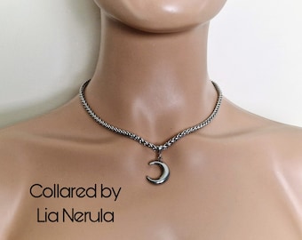 May include: A silver chain choker necklace with a crescent moon pendant. The necklace is on a mannequin's neck. The text "Collared by Lia Nerula" is below the necklace.