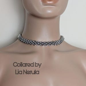 May include: A silver-colored chainmail choker necklace. The necklace is made of small, interlocking metal rings, creating a textured, layered design. The text "Collared by Lia Nerula" is displayed in a stylized font.