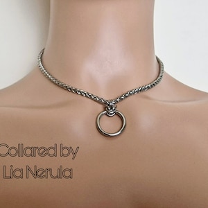 May include: A silver chain choker necklace with a large silver ring pendant. The necklace is on a model's neck. The text "Collared by Lia Nerula" is below the necklace.