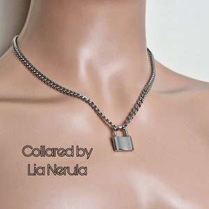 May include: A silver chain necklace with a silver padlock pendant. The necklace is on a mannequin's neck. The text "Collared by Lia Nerula" is visible below the necklace.