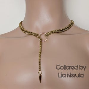 May include: A gold chain necklace with a large ring and a pointed pendant. The necklace is layered and has a delicate, feminine feel. The text "Collared by Lia Nerula" is visible in the image.