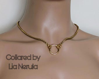 May include: A gold chain choker necklace with a round ring pendant. The necklace is on a model's neck. The text "Collared by Lia Nerula" is visible below the necklace.