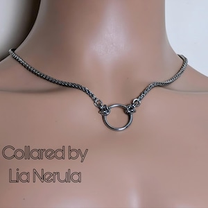 May include: A silver choker necklace with a circular pendant. The necklace has a woven chain design and a polished ring at the centre. The text "Collared by Lia Nerula" is displayed in a stylized font.