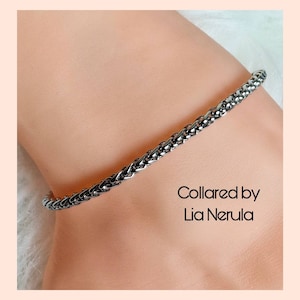 May include: A silver chain bracelet with a braided design. The bracelet is on a person's wrist. The text "Collared by Lia Nerula" is visible below the bracelet.