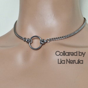 May include: A silver choker necklace with a large, round ring in the center. The necklace is made of a chain link design. The text "Collared by Lia Nerula" is visible below the necklace.