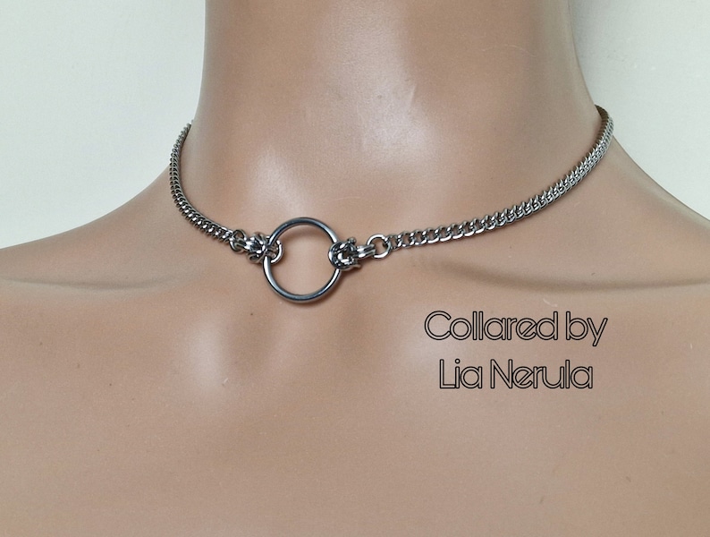May include: A silver choker necklace with a large, round ring in the center. The necklace is made of a chain link design. The text "Collared by Lia Nerula" is visible below the necklace.