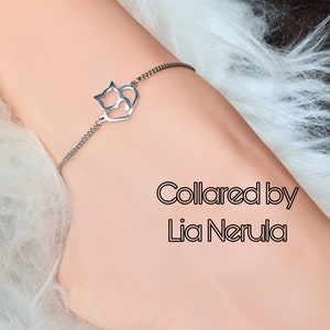 May include: A silver chain bracelet with a heart-shaped charm featuring three cats. The charm is cut out, creating a silhouette effect. The bracelet is on a person's wrist. The text "Collared by Lia Nerula" is visible below the bracelet.