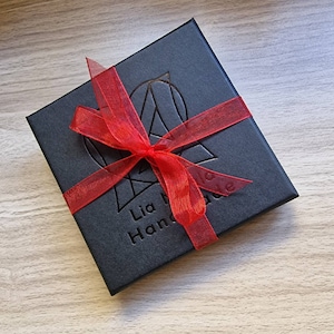 May include: A black gift box with a red ribbon tied around it. The box has the text "Lia Made Handmade" printed on it.