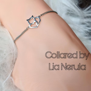 May include: A silver chain bracelet with a heart-shaped charm featuring three cats. The charm is made of metal and has a cutout design. The bracelet is on a white furry surface. The text "Collared by Lia Nerula" is visible below the bracelet.