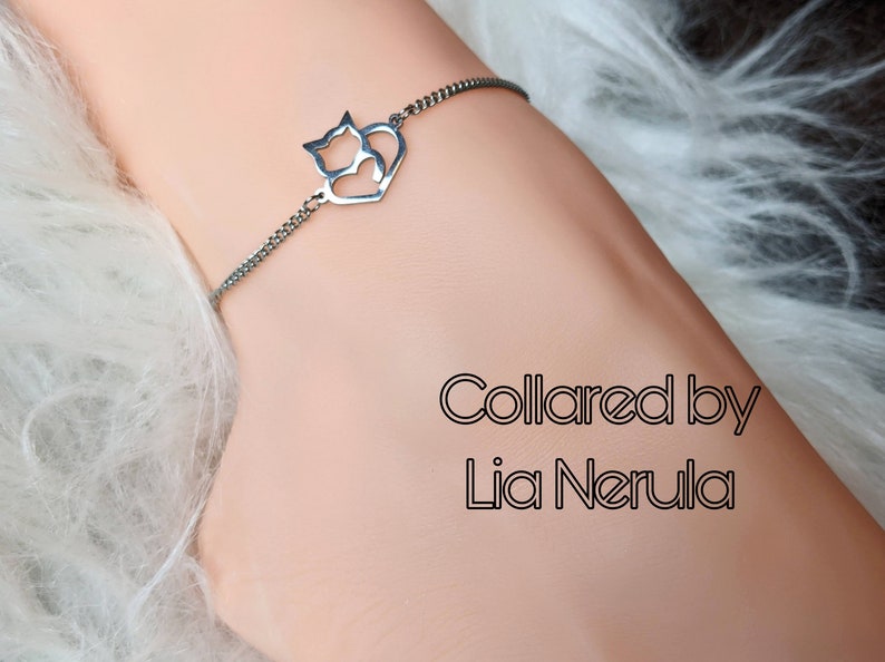 May include: A silver chain bracelet with a heart-shaped charm featuring three cats. The charm is made of metal and has a cutout design. The bracelet is on a white furry surface. The text "Collared by Lia Nerula" is visible below the bracelet.