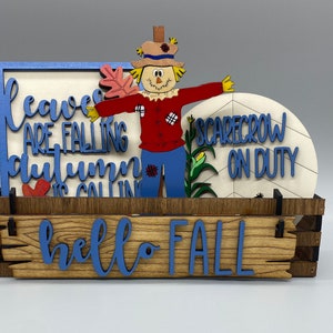 May include: A wooden box with a blue and white sign that says "leaves are falling, autumn is calling" and a smaller sign that says "hello fall". There is a scarecrow with a sign that says "scarecrow on duty" in front of the box.