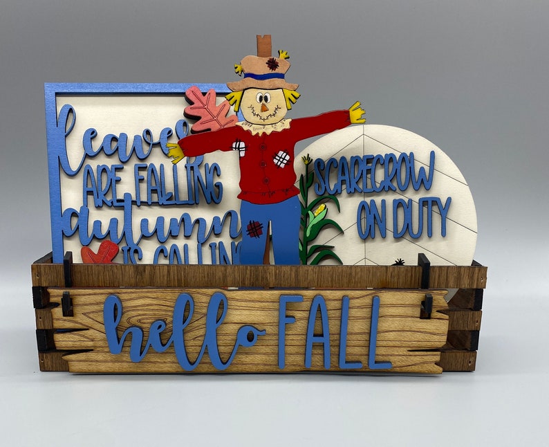 May include: A wooden box with a blue and white sign that says "leaves are falling, autumn is calling" and a smaller sign that says "hello fall". There is a scarecrow with a sign that says "scarecrow on duty" in front of the box.