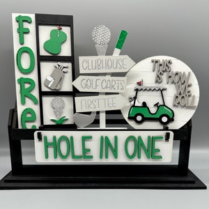 May include: A wooden sign with the words "FORE" and "HOLE IN ONE" in green letters. The sign also features a golf cart, golf clubs, and a sign that says "This is how we roll".