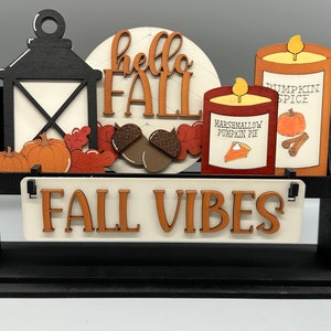 May include: A wooden tiered stand with a black base and black supports. The stand features a sign that reads "FALL VIBES" in brown letters. The top tier has a white circle with the words "hello FALL" in brown letters. There are two candles, one with a label that reads "MARSHMALLOW PUMPKIN PIE" and the other with a label that reads "PUMPKIN SPICE". There are also two pumpkins, two acorns, and some fall leaves.
