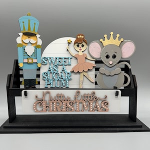 May include: A wooden Christmas decoration featuring a blue nutcracker, a ballerina, and a mouse wearing a crown. The words "Sweet as a Sugar Plum" and "Nutty Little Christmas" are also displayed. The decoration is set on a black base.