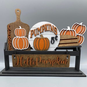 May include: A wooden stand with a sign that says "Pumpkins 5¢" and a smaller sign that says "Hello Pumpkin". There are also several wooden pumpkins on the stand.