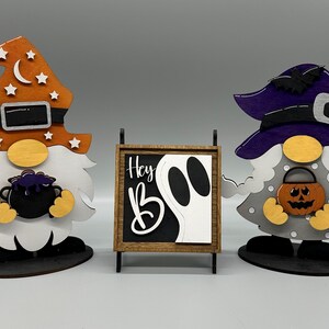 May include: Two Halloween gnomes, one with an orange hat and a cauldron, the other with a purple hat and a pumpkin. A wooden sign with the text "Hey Boo" and a ghost graphic.