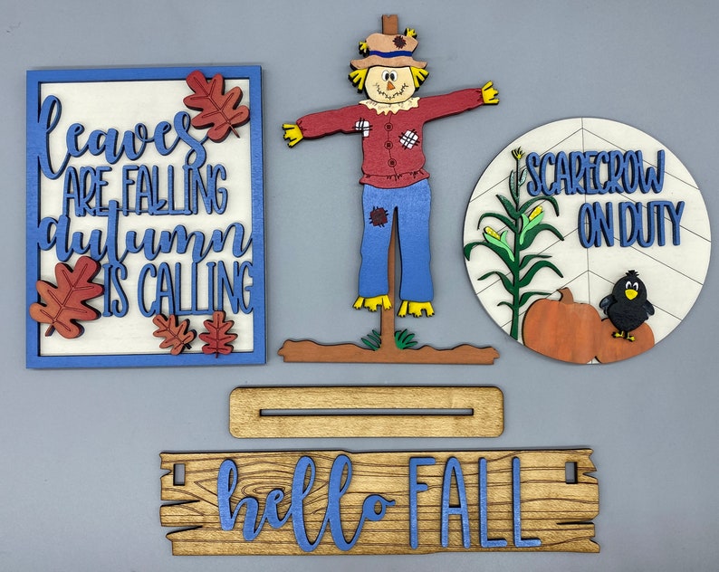 May include: A set of four wooden signs with a fall theme. The signs feature the text "leaves are falling autumn is calling", "hello fall", and "scarecrow on duty". The set also includes a wooden scarecrow figure.