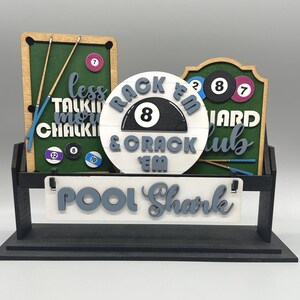 May include: A wooden pool-themed sign with a black base. The sign features a green felt-like background with pool balls, cues, and the text "Less Talking, More Chalking" and "Pool Shark". The center has "Rack 'Em & Crack 'Em".