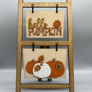 May include: Wooden easel with two signs. The top sign says "hello pumpkin" with a small orange pumpkin. The bottom sign has three pumpkins, two orange and one white, with leaves and a sunflower.