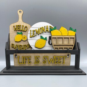 May include: Wooden sign with the words "Hello Sunshine" and "Life is Sweet" in yellow letters. The sign also features a round wooden sign with the words "Lemonade" in white letters and a basket of lemons.