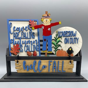 May include: A wooden sign with the text "Leaves are falling, autumn is calling" and a scarecrow with the text "Scarecrow on duty" on a black stand with a wooden sign that says "hello fall".