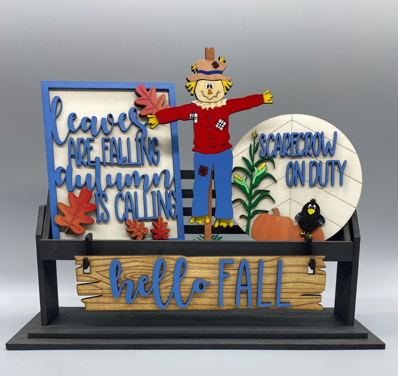May include: A wooden sign with the text "Leaves are falling, autumn is calling" and a scarecrow with the text "Scarecrow on duty" on a black stand with a wooden sign that says "hello fall".