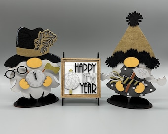 May include: Two wooden gnomes with black hats and white beards, one holding a clock and the other a sign that says "Kiss Me". A wooden sign in the middle says "Happy New Year".