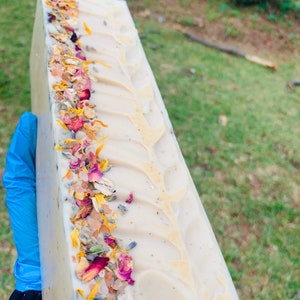 May include: A white bar of soap with a decorative swirl design. The soap is decorated with dried flowers, herbs, and pink Himalayan salt.