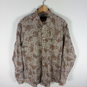 May include: A long-sleeve button-down shirt featuring a beige base with vertical pinstripes, a repeating brown paisley pattern, and small purple floral accents. The shirt includes a standard collar, a front chest pocket, and light-colored buttons.