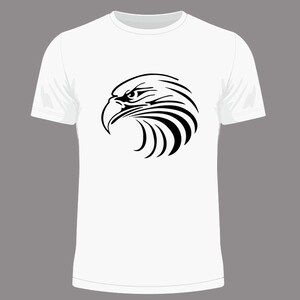 May include: White t-shirt featuring a black graphic of an eagle's head in profile. The eagle design is detailed with sharp lines, emphasizing the bird's beak and eye. The shirt has a classic crew neck and short sleeves.