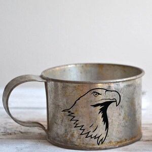 May include: A vintage metal mug with a handle, featuring a black graphic of an eagle's head. The mug has a distressed, aged appearance with visible wear and tear, suggesting it's an antique or a rustic-style item.