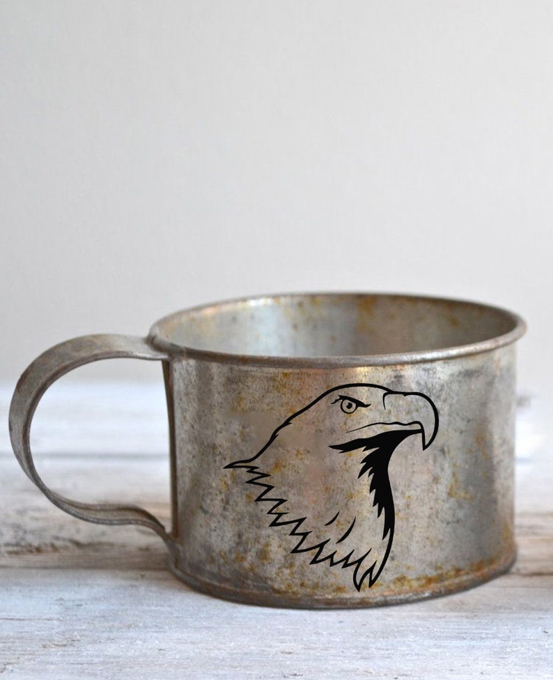 May include: A vintage metal mug with a handle, featuring a black graphic of an eagle's head. The mug has a distressed, aged appearance with visible wear and tear, suggesting it's an antique or a rustic-style item.