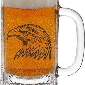 May include: A clear glass beer mug filled with amber-colored beer and a foamy white head. A black graphic of an eagle's head is on the front of the mug. The mug has a handle and is covered in condensation.