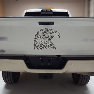 May include: A black and white graphic of an eagle's head is affixed to the tailgate of a white pickup truck. The truck has a tan bumper and black tires. The eagle graphic is a detailed line drawing.