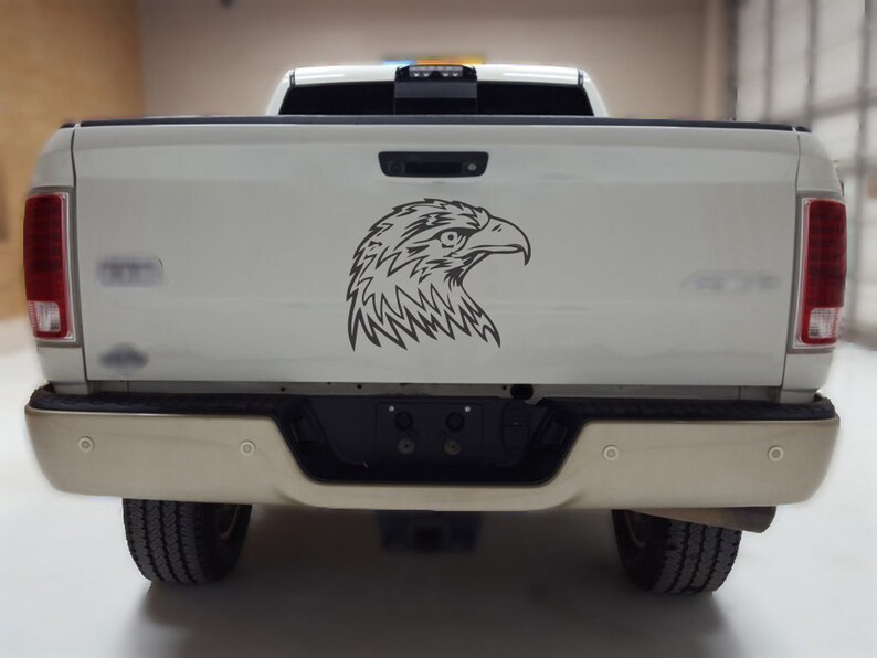 May include: A black and white graphic of an eagle's head is affixed to the tailgate of a white pickup truck. The truck has a tan bumper and black tires. The eagle graphic is a detailed line drawing.