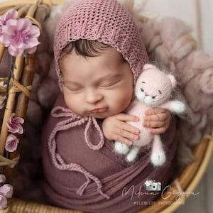 May include: A sleeping infant wrapped in a dusty rose fabric swaddle, wearing a matching knitted bonnet with ties. The infant holds a small, pale pink knitted cat toy. They are nestled inside a round, light-colored woven basket decorated with pink flowers.