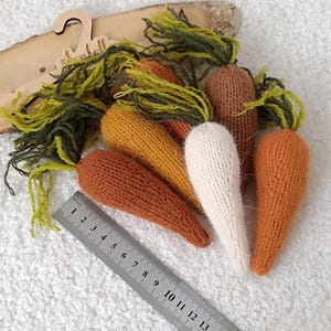 May include: A collection of handmade knitted carrots in various shades of orange, brown, and cream, with green yarn tops. A ruler is placed next to the carrots. The carrots are approximately 5 inches long.