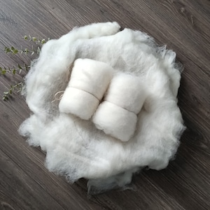 Könnte beinhalten: Zwei weiße, flauschige Garnrollen sind mit einer Schnur zusammengebunden und auf einer weißen, flauschigen Decke platziert. Die Decke liegt auf einer braunen Holzoberfläche.